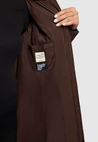 Jachetă izolată maro, parțial deschisă, care dezvăluie o căptușeală netedă, un eticheta și un buzunar pe partea stângă.