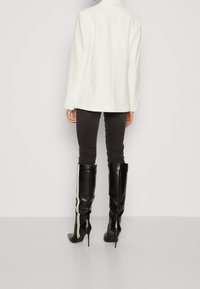 Person som bär en vit kavaj, mörka skinny-byxor och svarta knähöga klackstövlar med vita ränder, stående mot en enkel bakgrund.