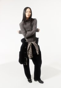 Person mit langen, glatten Haaren, die einen grauen Rollkragenpullover und schwarze Hosen tragen, mit einem karierten Hemd, das um die Taille gebunden ist, und einer schwarzen Jacke.