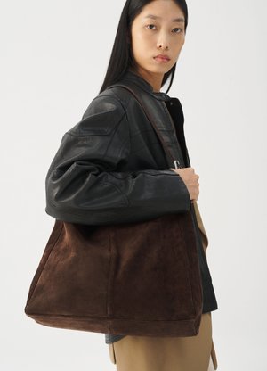 Woman wearing black leather jacket and beige trousers carries large brown suede tote bag over shoulder.