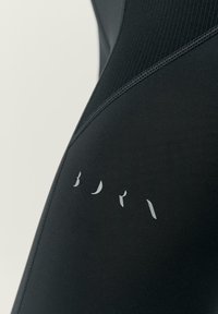 Fekete sportos ruházat, textúrázott mintával, letisztult dizájnnal és diszkrét szürke márkajelzéssel. Az anyag rugalmasnak és testhez simulónak tűnik.