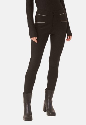 Mujer con pantalones negros ajustados con cuatro bolsillos delanteros con cremallera, una camiseta negra de manga larga y botines negros con tacón.