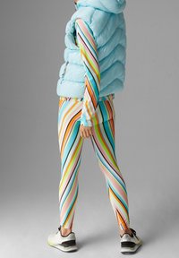 Světle modrá prošívaná vesta přes vícebarevné pruhované legíny s růžovými, oranžovými a zelenými pruhy na hladkém materiálu. Pod tím bílé tenisky.