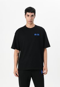 Camiseta negra de gran tamaño hecha de algodón, con un logo azul en el lado izquierdo del pecho. El diseño tiene un cuello redondo clásico y mangas cortas.