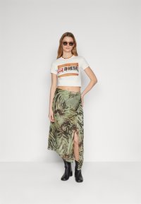 Tricou scurt crem cu un logo Diesel mare, de culoare maro. Fustă lungă verde cu un model de tigru și fante laterale. Bocanci negri până la gleznă. Ochelari de soare rotunzi.