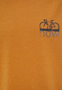 Tissu orange avec un petit motif de bicyclette bleu marine au-dessus du mot "maintenant" imprimé sur le côté supérieur gauche.