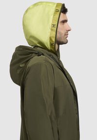 Olivgrüne Jacke mit einer kontrastierenden, leuchtend gelben Kapuze, ausgestattet mit einem Reißverschluss vorne und verstellbarem Zugband. Glatte Stoffoberfläche sichtbar.