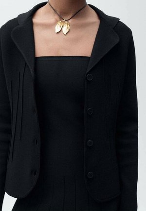Mujer con chaqueta negra texturizada y top sin tirantes, complementado con un collar con colgante de hoja dorada en un cordón negro fino.