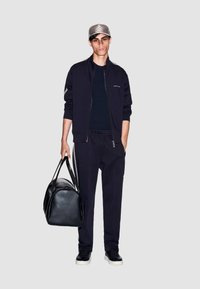 Zwart sportjack en broek gecombineerd met een zwarte leren duffeltas. Het model draagt een grijze pet, wat een stijlvolle, moderne uitstraling benadrukt.
