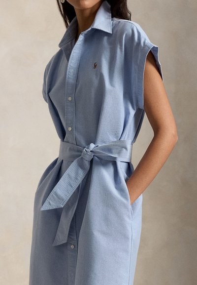 Γυναίκα με αμάνικο ανοιχτό μπλε φόρεμα με κουμπιά, γιακά και δεμένη ζώνη στη μέση, με το χέρι στην τσέπη, σε ουδέτερο φόντο.