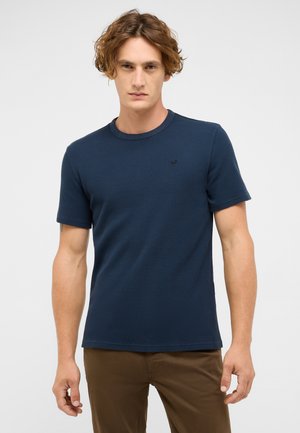Marineblaue Kurzarm-T-Shirt aus strukturiertem Stoff, mit rundem Ausschnitt und kleinem Logo-Akzent. Kombiniert mit braunen Hosen.