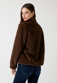 Giacca in pelliccia sintetica marrone con colletto alto, maniche lunghe e vestibilità rilassata. Texture morbida a contrasto con jeans di denim scuro, cuciture minime visibili.