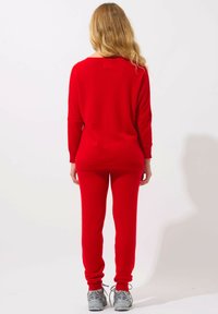 Roter Strickpullover und passende Hose, beide im lässigen Schnitt. Der Pullover hat weite Ärmel und einen Rundhalsausschnitt. Graue Sneakers werden getragen.