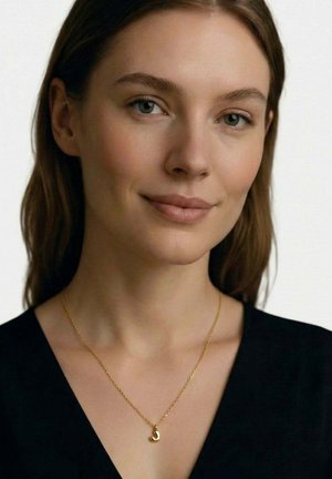 Jeune femme à la peau claire, cheveux bruns, yeux verts, portant un haut noir et un collier en or avec un petit pendentif incurvé.