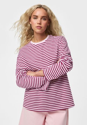 Jeune femme aux longs cheveux blonds portant un t-shirt à manches longues rayé rose et blanc et un pantalon rose clair, les bras croisés.