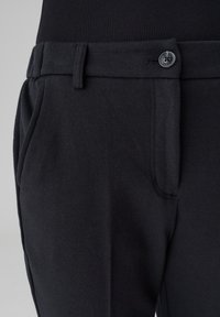 Gros plan sur un pantalon noir sur mesure avec taille, passants de ceinture, fermoir bouton et poche latérale porté par une personne vêtue d’un haut noir.