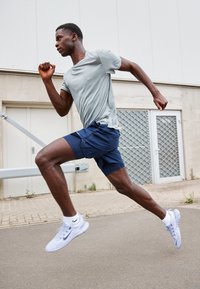 Atletisk mand i grå skjorte og blå shorts, der sprintet udendørs på fortovet nær en industribygning.