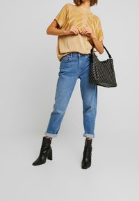Mujer vestida con una blusa dorada con estampado, jeans azules remangados, botas de tacón negras a nivel del tobillo, sosteniendo un bolso negro texturizado.