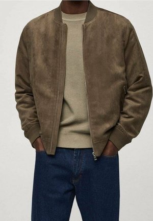 Mand iført en brun ruskind bomberjakke over en beige strikket sweater og blå jeans med hænderne i jakkelommerne.