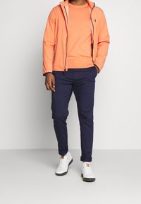 Lätt orange jacka med huva, marinblå byxor och vita sneakers. Har dragkedja framtill, sidofickor och uppvikta manschetter på byxorna.