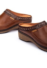 Sabots en cuir marron avec une bande décorative cousue, un intérieur rembourré et une semelle compensée texturée. Étiquette de marque visible sur le côté.