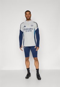 Grå og marineblå sportsoutfit med lange ærmer og lynlås krave. "Emirates Fly Better" logo på brystet, shorts i matchende ærmer farve og sorte sko.