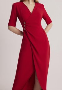 Robe midi portefeuille rouge avec trois boutons décoratifs sur le côté, manches courtes et une fente frontale, portée par une personne les mains sur les hanches.