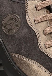Zapatilla deportiva en marrón y gris, con gamuza y cuero texturizado, un parche con el logo y cordones de tela. Diseño de suela curvada.