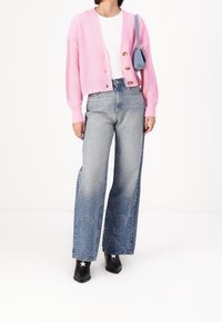 Kvinna i en rosa knäppt cardigan, vit topp, mönstrade vida jeans, svarta stövlar med vita stjärnor och en liten blå axelremsväska.