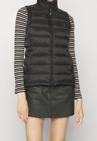 Gilet nero in piuma con collo alto e zip, indossato sopra una maglietta a righe a maniche lunghe. Combinato con una gonna nera con orlo sfrangiato.