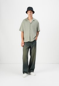 Camisa de manga corta de color verde claro con un patrón y cuello, combinada con unos vaqueros oscuros holgados y zapatillas blancas. Se lleva un sombrero tipo cubo negro.