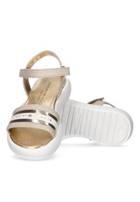 Sandalias beige con una correa metálica dorada y suela blanca. Presenta una banda con logo y una textura suave. Correa en el tobillo para un ajuste ajustable.