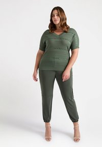 Olijfgroene gebreide top met korte mouwen en een V-hals, met textuurpatronen. Gecombineerd met olijfgroene joggers en neutrale sandalen.