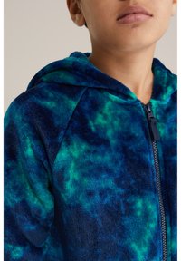 Huvtröja i fleece i blått med ett grönt tie-dye mönster. Har dragkedja och mjukt, texturerat tyg för komfort.