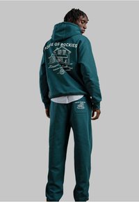 Felpa con cappuccio color teal e pantaloni jogging coordinati presentano testo e grafiche bianche, inclusa una grafica di una casa. Materiale in pile con vestibilità rilassata e tasche.