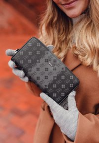 Gray wallet featuring a textured pattern of paws and a logo, with a zipper closure. Held with gray gloves against a brick background.
