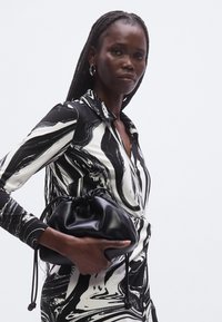 Svart lærpose med snøring holdt i hånden. Modellen har på seg en tettsittende svart- og hvitmønstret kjole med krage.