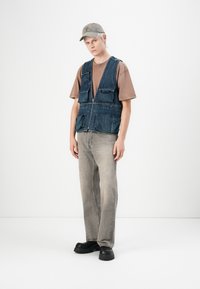 Vesta din denim cu multiple buzunare, închidere cu fermoar, purtată peste un tricou maro. Blugi gri deschis și pantofi negri masivi completează ținuta.