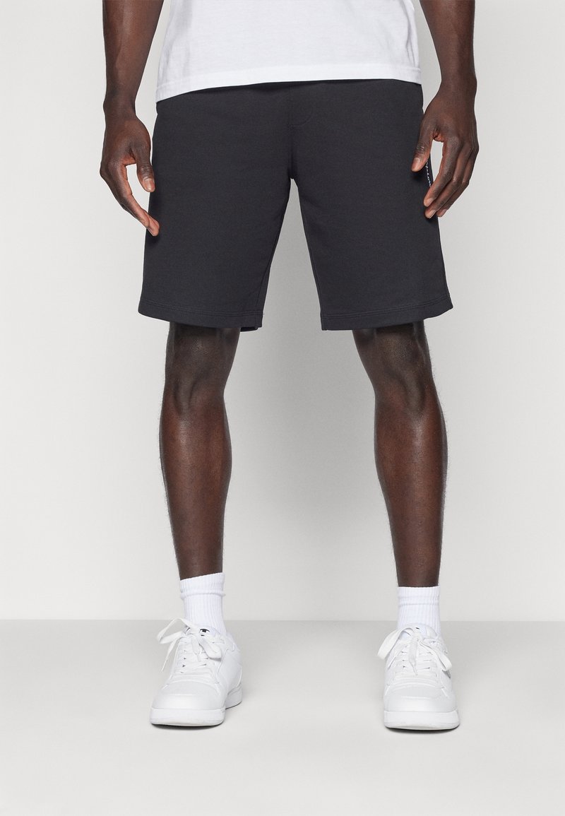 Svarta bomullsshorts med rak skärning, har förstärkt midja, sidofickor och en mjuk textur; ihop med vita sneakers och strumpor.