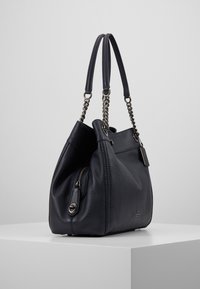 Sac à bandoulière en cuir noir avec double chaîne et poignées en cuir, poche latérale zippée, et ferrures argentées, posé sur un piédestal blanc.