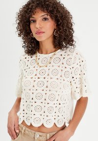 Top au crochet en crème avec motifs circulaires, manches courtes et ourlet festonné. Le tissu est léger et texturé, améliorant la respirabilité.