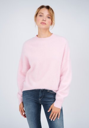 Frau mit heller Haut und blondem Haar, die einen weichen rosa Pullover und blaue Jeans trägt, steht vor einem einfarbigen hellen Hintergrund.