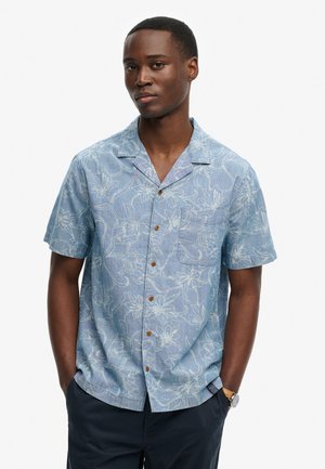 Młody mężczyzna w jasnoniebieskiej koszuli z krótkim rękawem z białym kwiatowym wzorem i ciemnych spodniach, ręce w kieszeniach.