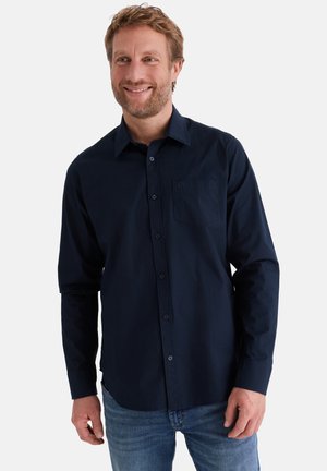 Lächelnder Mann mit hellbraunen Haaren, der ein dunkelblaues, langärmliges Hemd mit Knöpfen und blaue Jeans vor einem schlichten Hintergrund trägt.