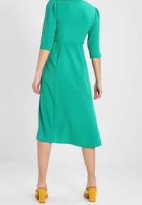 Green, knee-length dress with three-quarter sleeves and button accents at cuffs. Smooth fabric with a natural drape, paired with yellow block heels.