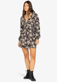 Vestido negro con flores de manga larga, escote en V, cintura ceñida y falda acampanada, con patrones florales en crema y dorado. Combinado con botas negras.