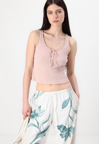 Light pink cropped tank top with a ribbed texture, featuring a lace trim and front tie closure. Paired with white pants adorned with teal floral print.