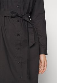 Robe chemisier noire en tissu lisse, avec une taille ceinturée, des manches longues, une poche poitrine unique et des boutons noirs.