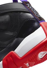 Gros plan d'une chaussure Air Jordan noire et rouge montrant le cuir texturé, la languette violette avec le label "JORDAN" et la semelle rouge à motifs.