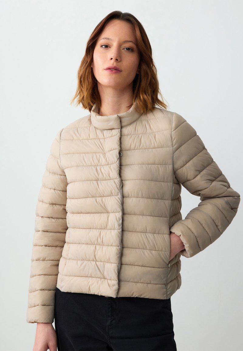 Giacca trapuntata beige con design a righe orizzontali, colletto alto e chiusura a bottoni. Tessuto liscio, finitura leggermente lucida e tasche laterali.